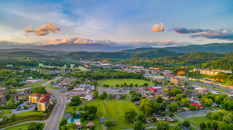Aerial view of Pigeon Forge, Tennessee, nestled in a valley surrounded by lush green hills and the Smoky Mountains under a partly cloudy sky at dusk.